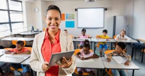 Female teacher standing with device in classroom